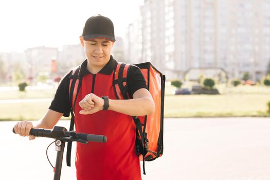 Positive deliveryman worker employee deliver online order client customer. Smiling man courier food delivery with red thermal backpack walks street with electric scooter.