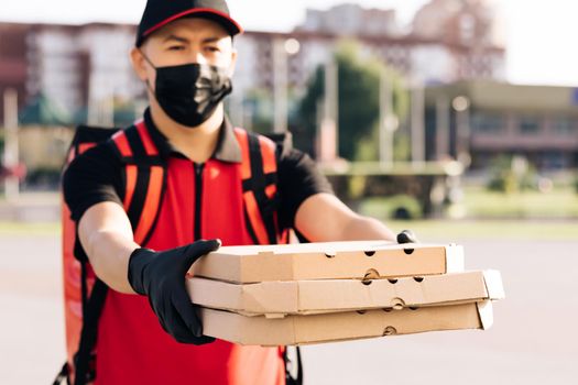 Portrait shot of handsome caucasian deliveryman in gloves and medical mask standing at street, holding and handing carton boxes. Male courier giving pizza.