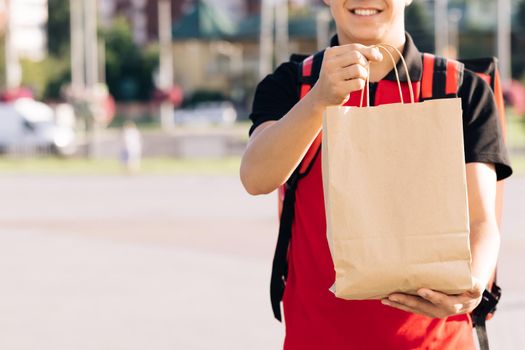 Close up portrait of positive young man courier person. Delivery service door to door. Happy delivery worker holding packet with food looking at the camera and smiling.