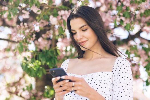 Portrait of a beautiful woman using a smartphone in the park on a background of sakura trees. Young beautiful girl browsing the Internet using social networks.