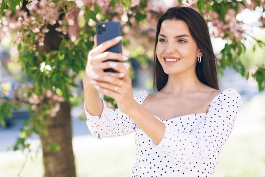 Smiling woman having video call on smartphone in city, friendly woman waving hand at camera, pretty woman video chatting at phone, stylish blogger making video on phone outdoor.