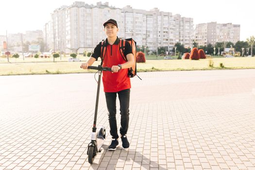 Smiling asian man courier food delivery with red thermal backpack walks street with electric scooter. Positive deliveryman worker employee deliver online order client customer.