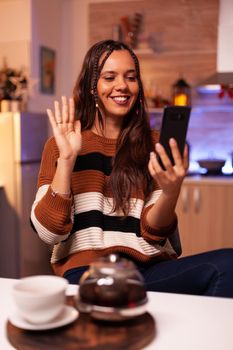 Caucasian person using video call conference technology on online internet, holding smartphone and cup of winter drink in decorated kitchen for holiday. Young cheerful woman being social