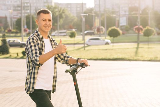 Portrait shot of caucasian hipster man leaning on electric scooter looking at camera and showing thumb up. Hipster man on vehicle outdoors. Eco-friendly modern urban transport.