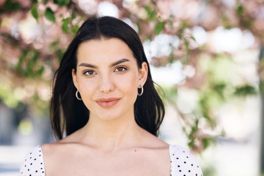 Perfect model with creative vivid makeup and pink lipstick on lips and hairstyle posing outside happy looking at camera. Spring blossom of sakura tree in city park.