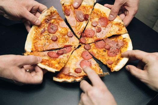 Group of hungry friends sitting at desk and sharing delicious lunch on table background. Male hands taking slices of pizza with cheese, tomatoes and ham from food delivery.