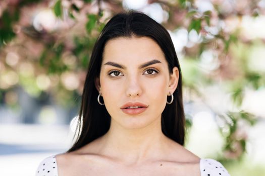 Front closeup portrait of young mixed race beauty american woman with long hair style standing on street near sakura trees, looking friendly at camera. Human face, ideal natural make up skin concept