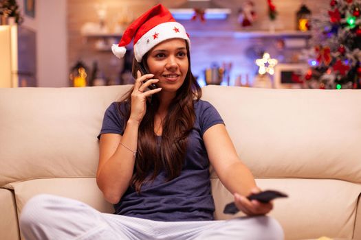 Portrait of woman talking at phone with friend while watching winter movie on television celebrating christmastime in x-mas decorated kitchen. Girl with santa hat enjoying winter season