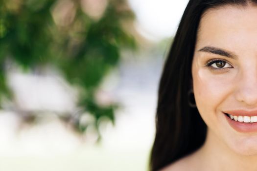 Close up half face portrait smiling attractive woman model looking at camera, happy beautiful 20s brunette lady pretty face dental smile posing stand alone at park on a background of sakura trees.