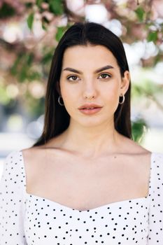 Vertical portrait of young beauty american woman with long hair style standing on street near sakura trees, looking friendly at camera. Human face, ideal natural make up skin concept