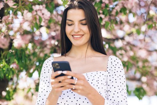 Smiling woman walks down the central park city street and uses her phone. Pretty summer woman in white dress walks down the street looking at her mobile phone.