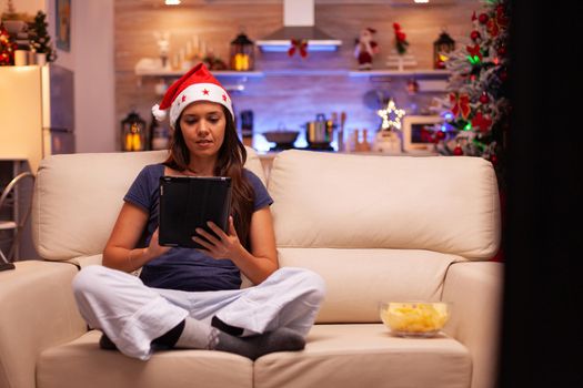 Adult woman wearing santa hat sitting in lotus position while browsing on social media using tablet during christmastime in xmas decorated kitchen. Caucasian female celebrating christmas holiday