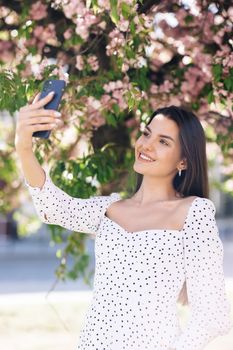 Smiling woman in summer white dress taking selfie self portrait photos on smartphone. Model posing on park sakura trees background. Female showing positive face emotions