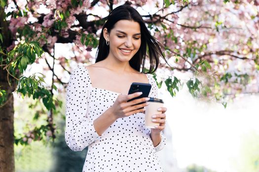 Young woman wearing white dress walking in the city center, using smartphone and drinking coffee. Communication, online shopping, social network concept.