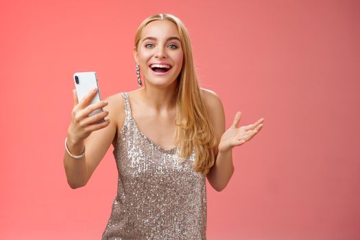 Astonished happy charming blond girl in silver glittering stylish dress holding smartphone amazed liking awesome result edit photo app smiling wondered amused, standing red background.