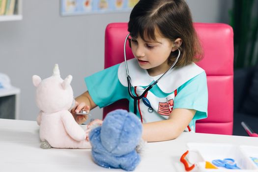 Cute little child girl wear medical clothing playing game as vet doctor holding stethoscope listening sick toy. Adorable preschool child pretending doctor treat toy patient.