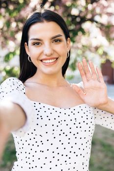 Vertical photo of smiling woman having video call on smartphone in city, friendly woman waving hand at camera, pretty woman video chatting at phone, stylish blogger making video on phone outdoor.