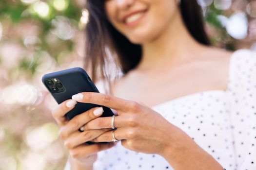 Cheerful girl with smartphone in park on a background of sakura trees. Smiling lady holding cellphone in hands outside. Happy hipster woman typing by mobile phone outdoors