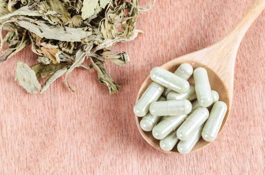 Pile of herbal medicine capsules in wooden spoon with dried leaves on wooden background.