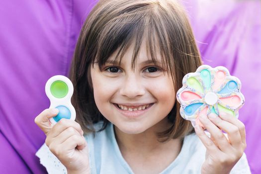 Portrait little girl with a modern popit toy. Colorful and bright pop it toy. Simple dimple. The child is sitting on a easy chair with colorful trendy antistress sensory toy pop it and simple dimple.