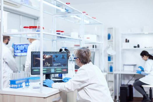 Elderly aged medical chemist working on powerful computer in modern facility. Senior scientist in pharmaceuticals laboratory doing genetic research wearing lab coat with team in the background.
