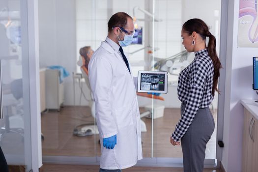 Dentist with face mask showing patient dental x-ray in stomatology specialist reception. Explaining diagnosis during appointment, senior man sitting in dental chair.