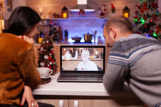 Happy family talking with remote grandmother during online videocall meeting teleconference sitting at table in xmas decorated kitchen. Joyful couple celebrating christmas holiday