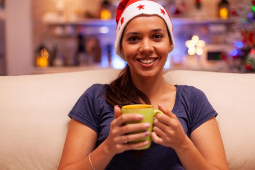 Portrait of woman wearing red santa hat holding cup of coffee in hands enjoying winter season relaxing on couch in xmas decorated kitchen. Adult celebrating christmas holiday