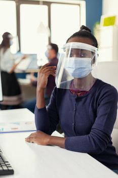 Entrepreneur sitting at her workplace wearing face mask against coronavirus. Multiethnic business team working in financial company respecting social distance during global pandemic.