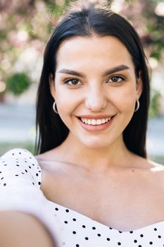 Young smiling woman in summer white dress taking selfie self portrait photos on smartphone. Model posing on park sakura trees background. Female showing positive face emotions