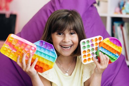 Portrait little girl with a modern popit toy. The child is sitting on a easy chair with colorful trendy antistress sensory toy pop it and simple dimple. Colorful and bright pop it toy. Simple dimple