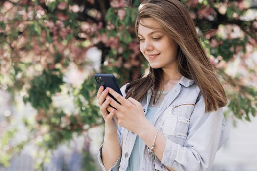 Portrait of happy hipster woman typing by mobile phone outdoors. Cheerful girl walking with smartphone in urban background. Smiling lady holding cellphone in hands outside.