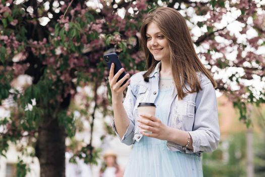 Beautiful Woman with charming Smile walking in the Park and Using Mobile Phone Wearing Stylish Outfit. Girl Using Smartphone and Drinking Coffee. Communication, online shopping, social network concept
