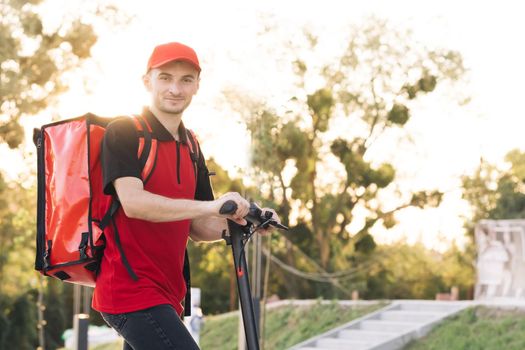 Young student works in a delivery service, with a red backpack and baseball cap, rides an electric scooter along the embankment at sunset.