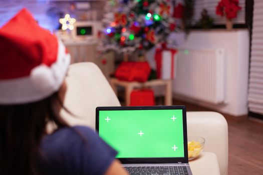 Caucasian female looking at mock up green screen chroma key laptop with isolated display resting on couch in xmas decorated kitchen. Woman enjoying winter season celebrating christmas holiday