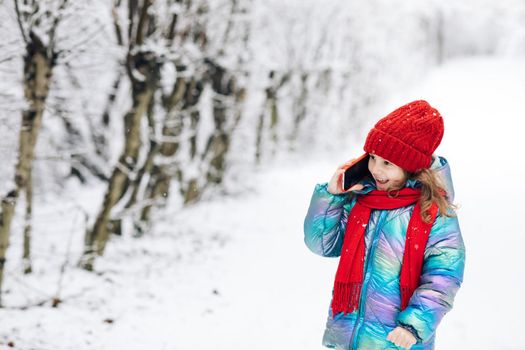 Pretty little child girl with curly hair in winter clothes talking on mobile phone smiling standing on the winter park near Christmas trees. Little child girl having conversation on her mobile phone