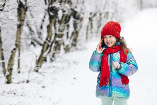 Young beautiful child girl in winter hat talking emotionally and surprised talking on cell phone. Young curly girl talking on her mobile phone while walking in the winter city street.