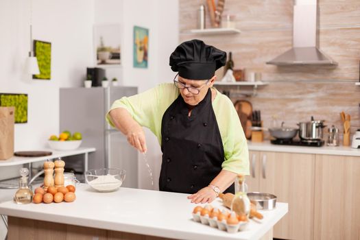 Woman is sifting flour on wooden table in modern kitchen. Happy elderly chef with uniform sprinkling, sieving sifting raw ingredients by hand baking homemade pizza.