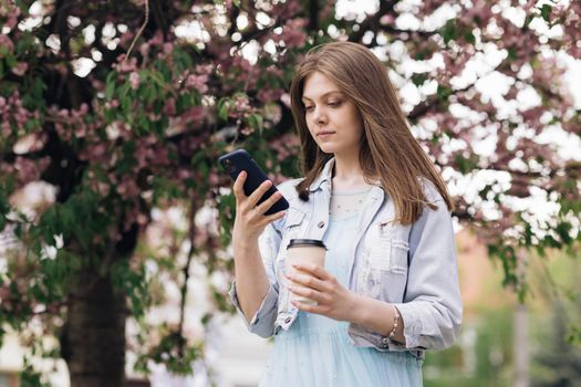 Young woman wearing white dress walking in the city center, using smartphone and drinking coffee. Communication, online shopping, social network concept.