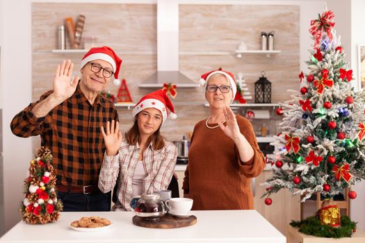 Pov of family wearing santa hats greeting friends during online videocall meeting conference enjoying winter season. Grandaprarents standing at table in xmas decorated culinary kitchen. Christmas call