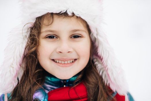 Funny cute girl laughing outdoors in winter day. Pretty little girl smiling with long hair in red hat and scarf standing in the white snowy park or forest at winter time