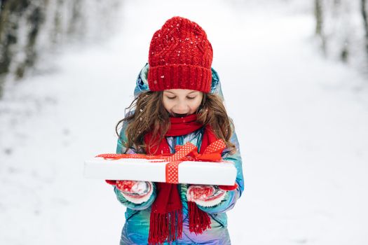 Child holding gift box and surprise face. Child in red hat with Christmas gift box in snow. Winter outdoor fun. Kid play in snowy park