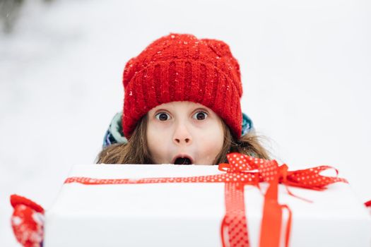 Smiling cute girl 6-7 years holding in hands birthday present in festive package on winter background. Portrait of happy girl peeking out of giftbox on Christmas day