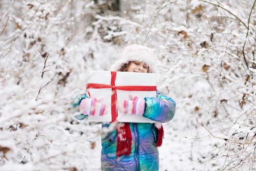 Happy child girl with Christmas gift on a winter walk in nature. Little girl enjoying the holidays. Portrait kid with gift box on winter background.