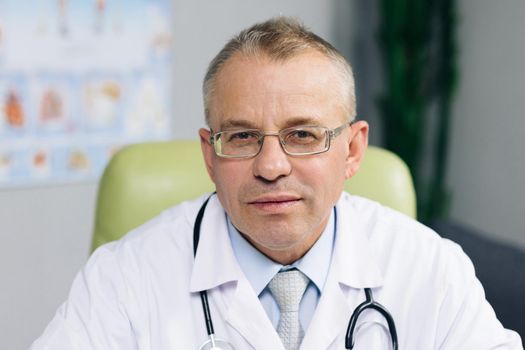 General practitioner looking at camera, posing in office. Portrait of confident old mature male head doctor physician in white medical uniform in glasses sitting at workplace.