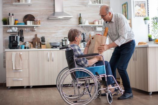 Senior man helping his wife by taking grocery paper bag from her. Mature people with fresh vegetables from market. Living with disabled person with walking disabilities