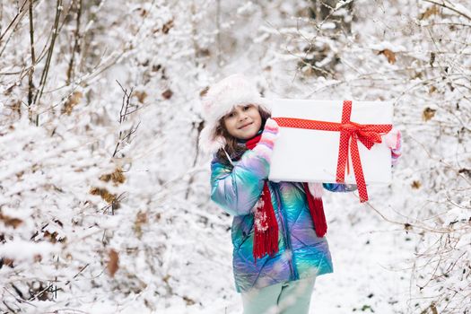 Child with Christmas presents and gifts in snow. Winter outdoor fun. Kid play in snowy park on Xmas eve. Happy Little caucasian girl smile and holding gift box.