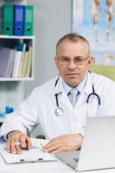 General practitioner looking at camera, posing in office. Confident old mature male head doctor physician in white medical uniform in glasses sitting at workplace.