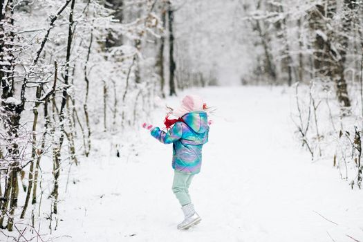 Child girl in winter playing in snow into park outdoor. Kid shakes snows off with fir trees. Winter time happiness concept. Snowing weather