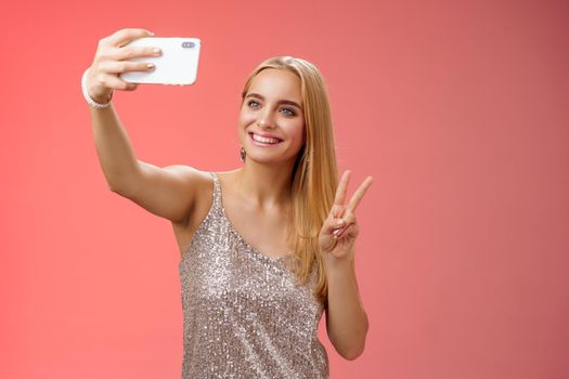 Attractive feminine tender young blond girl 25s in silver stylish dress taking selfie extend arm up show peace gesture smartphone display record video greeting internet fans, standing red background.